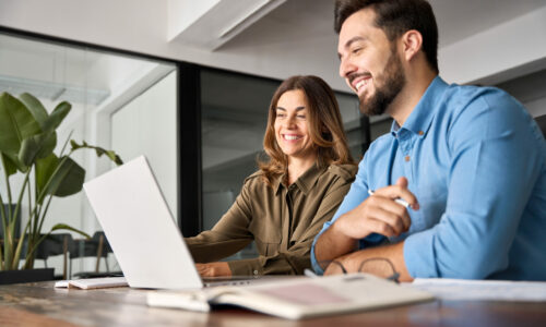 Two smiling professional business people talking using laptop computer working in office. Happy colleagues or entrepreneurs team man and woman discussing corporate technology at workplace.