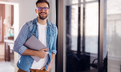 Portrait of a young modern businessman standing holding a laptop and looking at the camera with a happy smile.