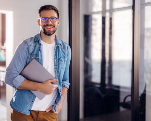 Portrait of a young modern businessman standing holding a laptop and looking at the camera with a happy smile.