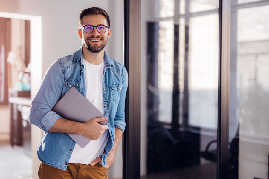 Portrait of a young modern businessman standing holding a laptop and looking at the camera with a happy smile.