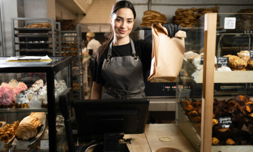 baker woman standing with fresh bread and pastry at bakery. Young woman in her bake shop looking at camera. Satisfied baker with breads in background. Girl owner bakery shop