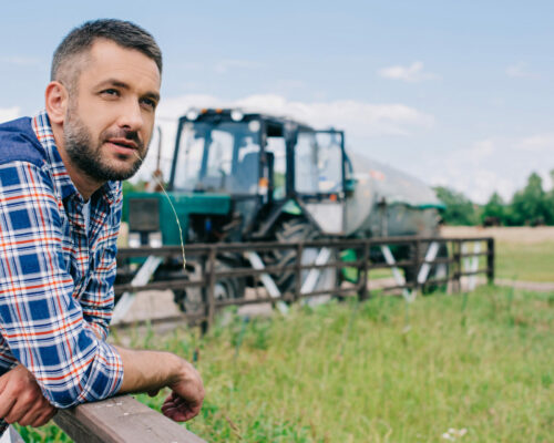 handsome middle aged farmer leaning at fence and looking away at farm