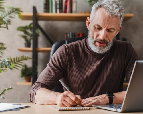 Middle aged businessman in casual clothes making notes in notepad while sitting in his office